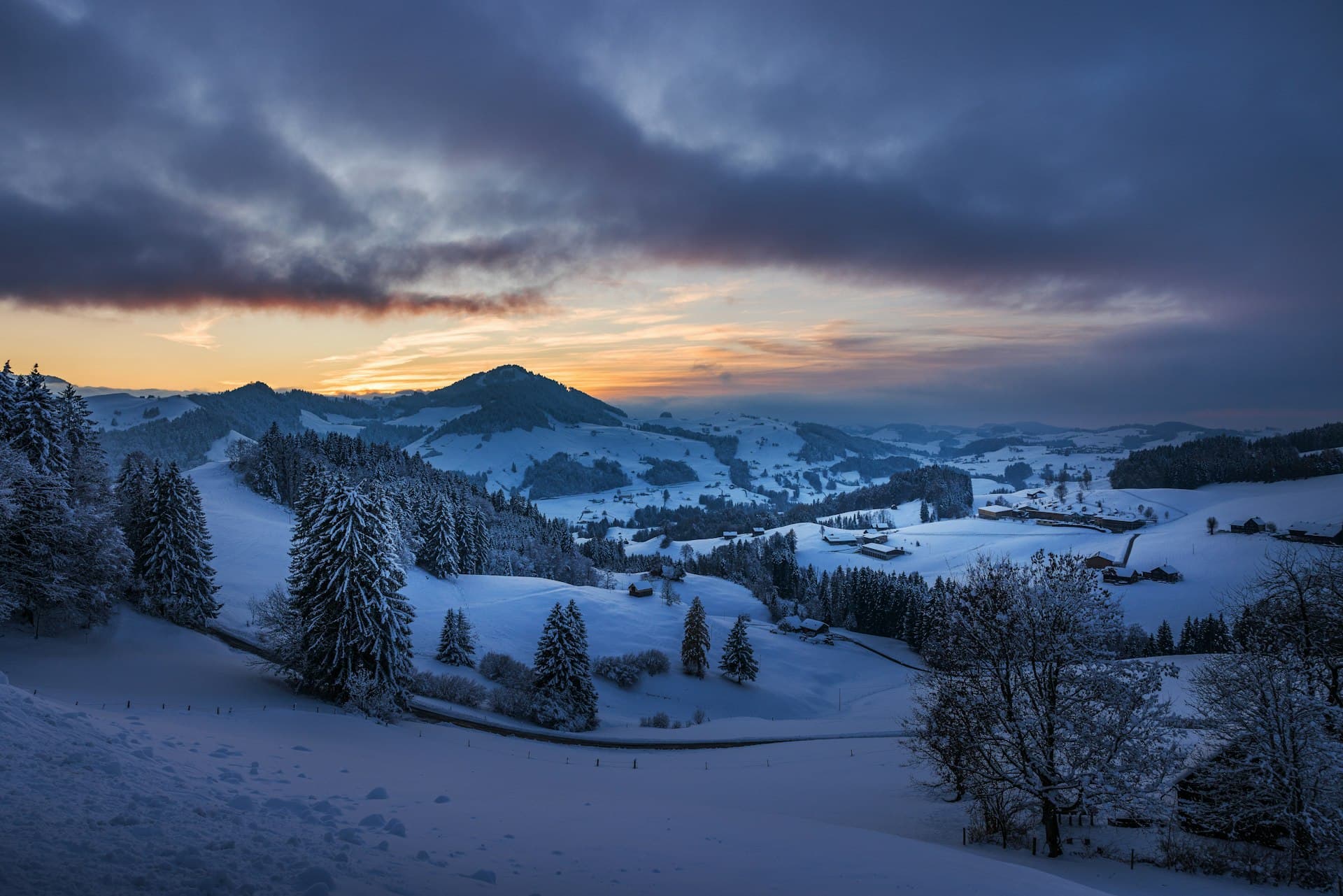 A sweeping alpine landscape at dusk beneath dramatic, brooding clouds