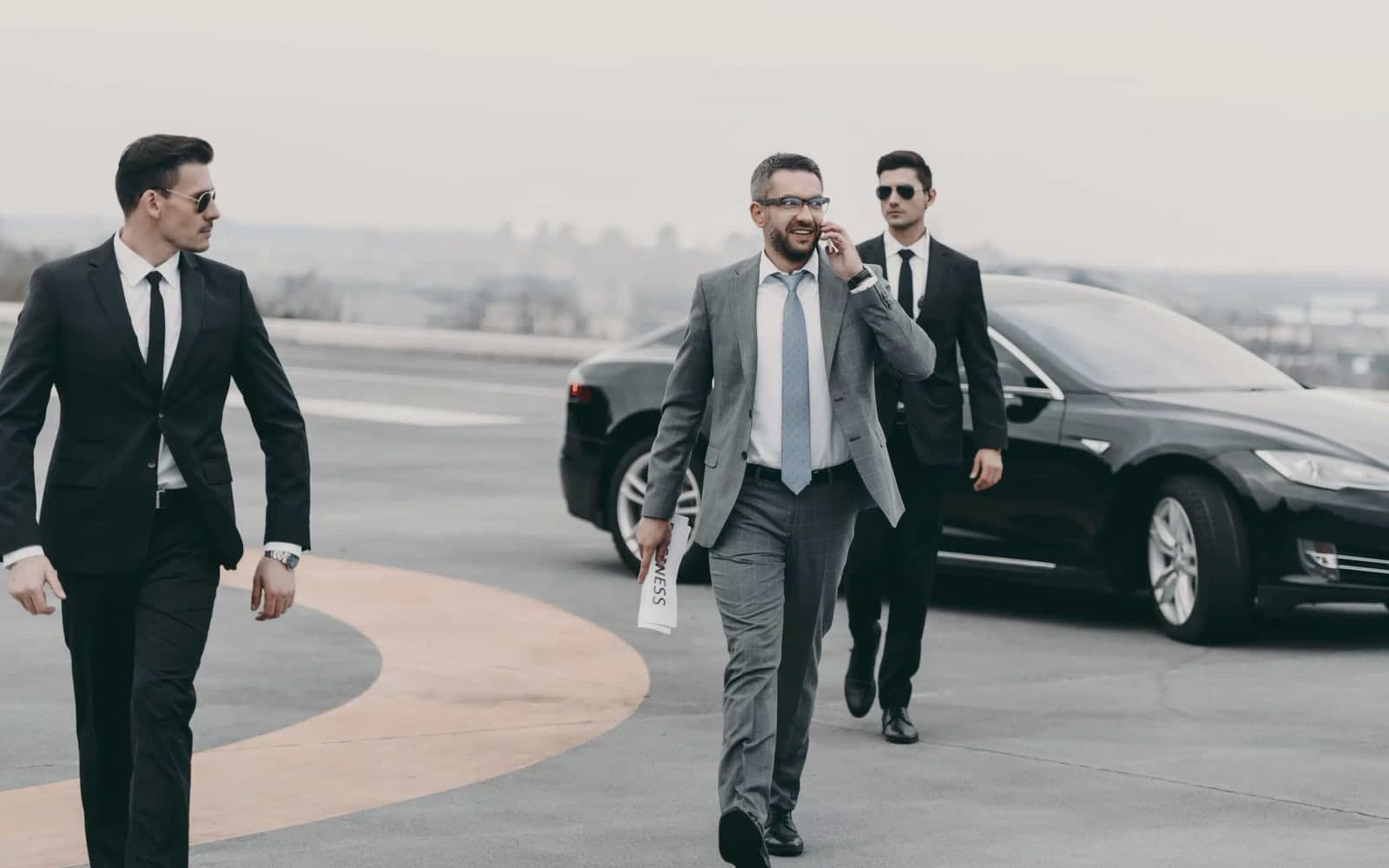 Close-protection personnel in suits coordinating a principal movement beside a black executive vehicle